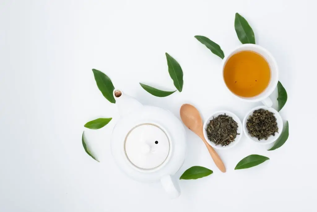 From above of cup with green tea near dry and fresh leaves with ceramic teapot on white table